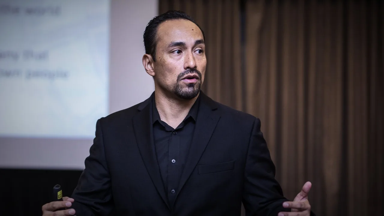 Juan P. Ortega presenting on stage in 2018, gesturing toward the audience in a dark suit with slides behind him